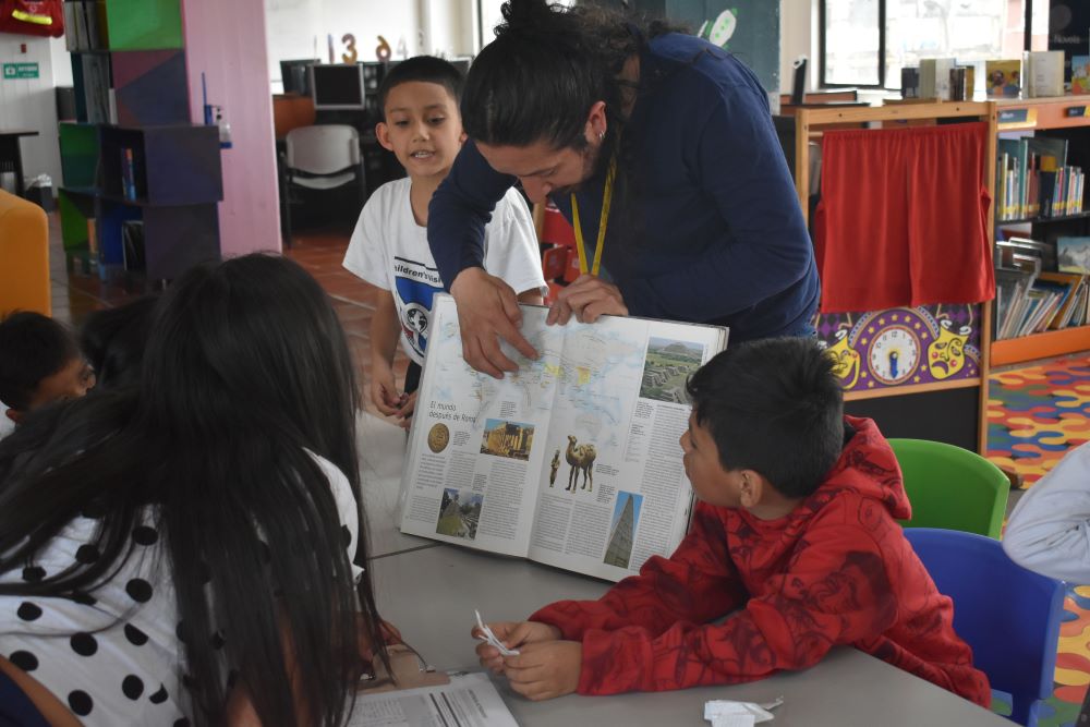 niños y niñas atentos a la lectura que esta realizando la profesora mientras les enseña un libro ilustrado.