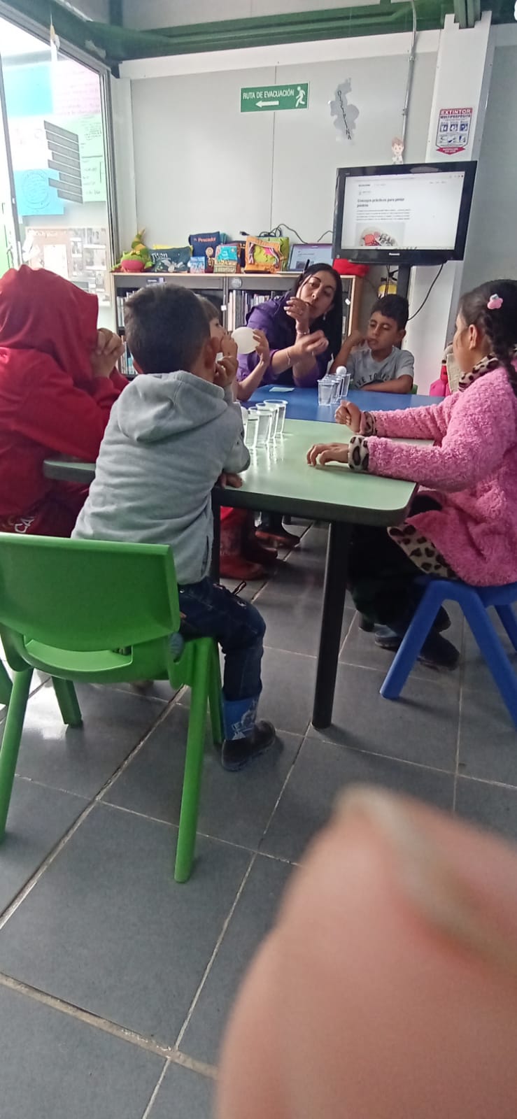 niñas y niños en medio de un taller en la biblioteca, sobre la mesa hay vasos de plástico llenos de agua