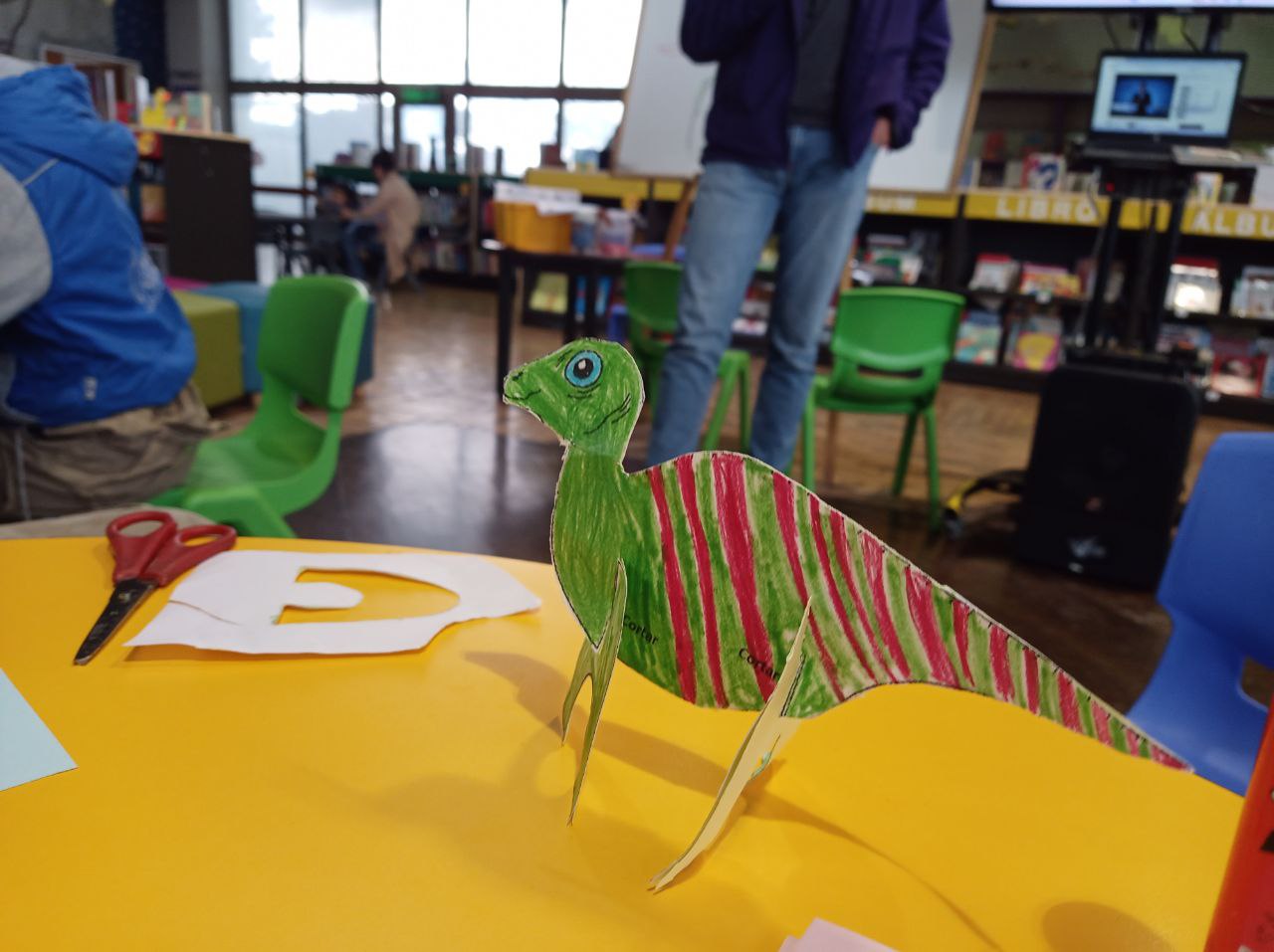 Este es un dinosaurio de papel hecho a mano en un aula. El dinosaurio, coloreado en tonos verdes y rojos, se encuentra sobre una mesa amarilla. Junto a él, hay unas tijeras rojas y negras, así como recortes de papel blanco. En el fondo, se ven sillas verdes y azules, estanterías con libros y una persona parada.
