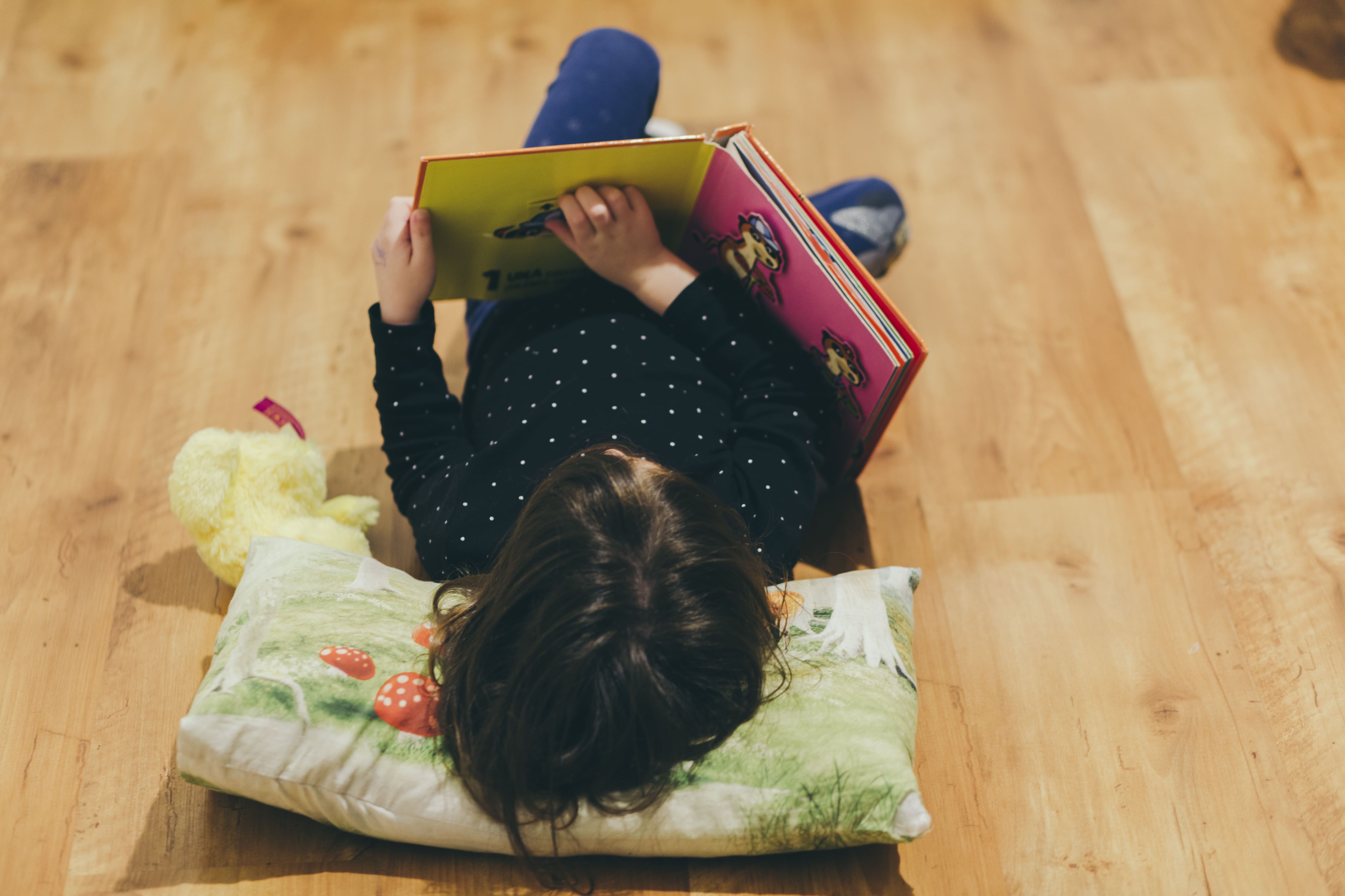 una niña acostada sobre una almohada en el piso leyendo un libro ilustrado.