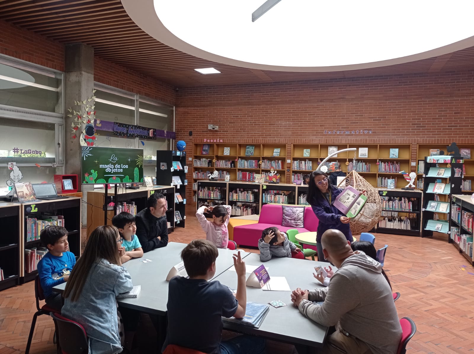 niños y niñas acompañados de sus padres al rededor de una mesa mientras leen en voz alta junto a la profesora.