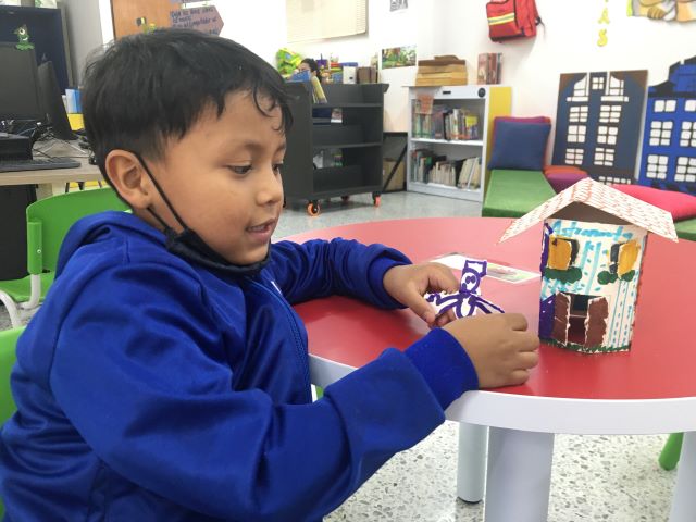 niño pequeño vestido de azul jugando con una casa y un muñeco de papel creados por él.