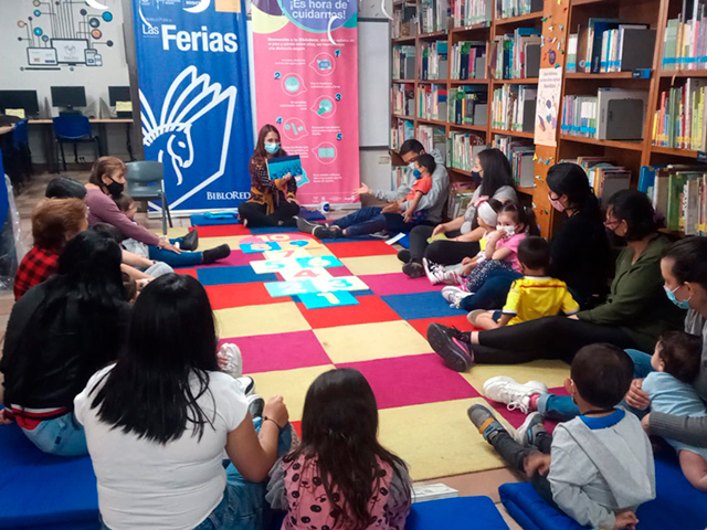 niños y niñas acompañados de sus padres sentados en círculo leyendo un libro en voz alta.