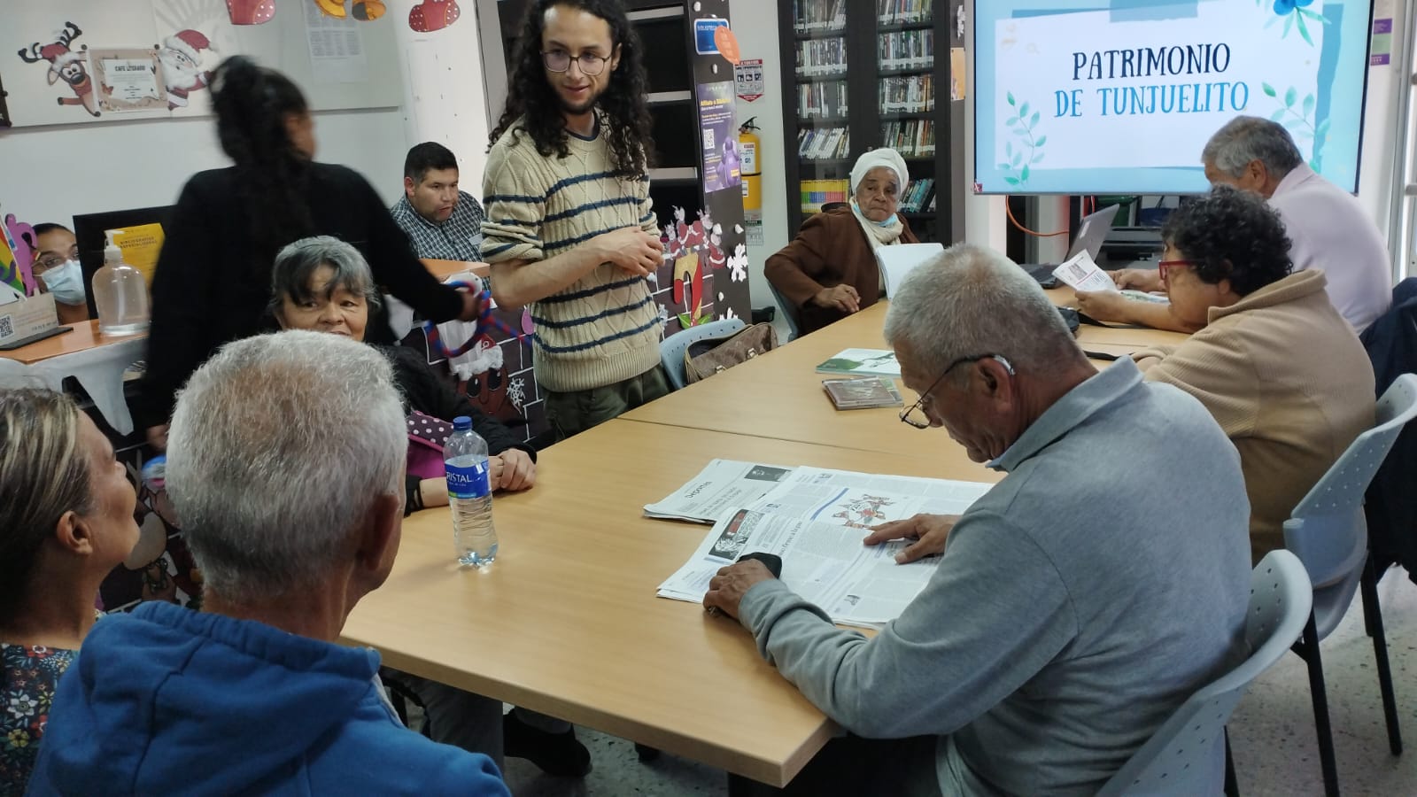 adultos mayores sentados en mesa redonda frente al docente mientras dialogan sobre un libro. 