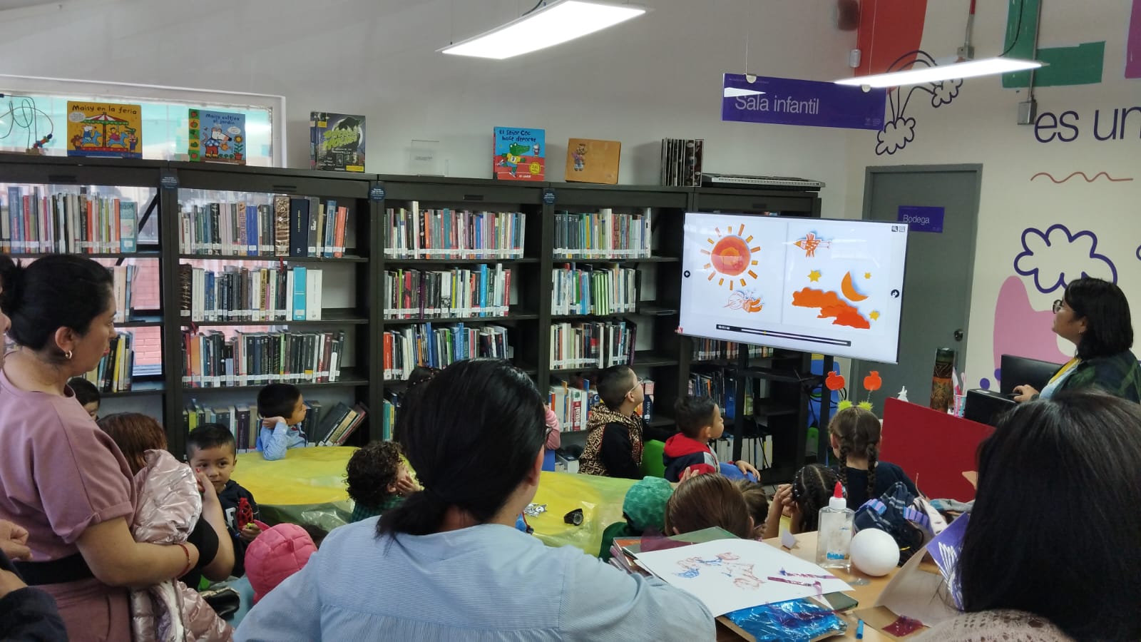 niños y niñas acompañados por sus cuidadores observando las páginas de un libro digital.