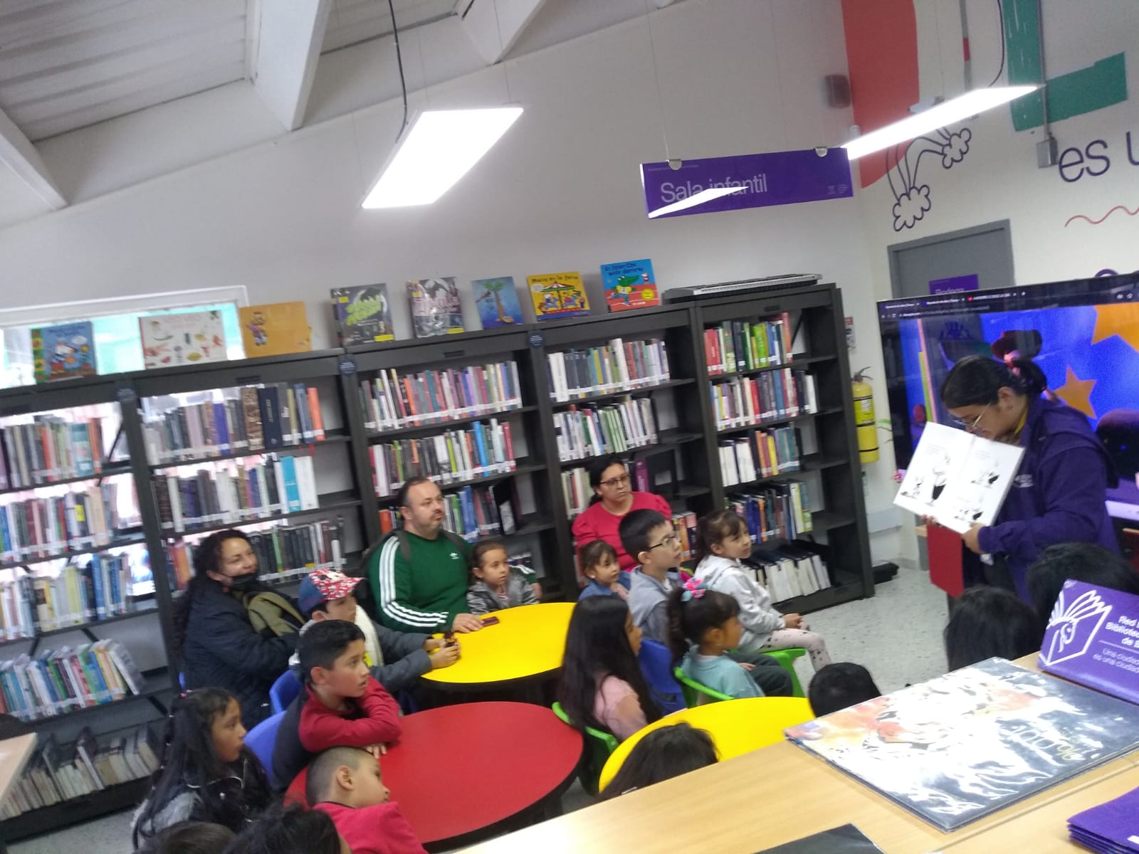 niños y niñas reunidos al rededor de la profesora escuchándola leer un libro en voz alta mientras disfrutan de las ilustraciones de mismo
