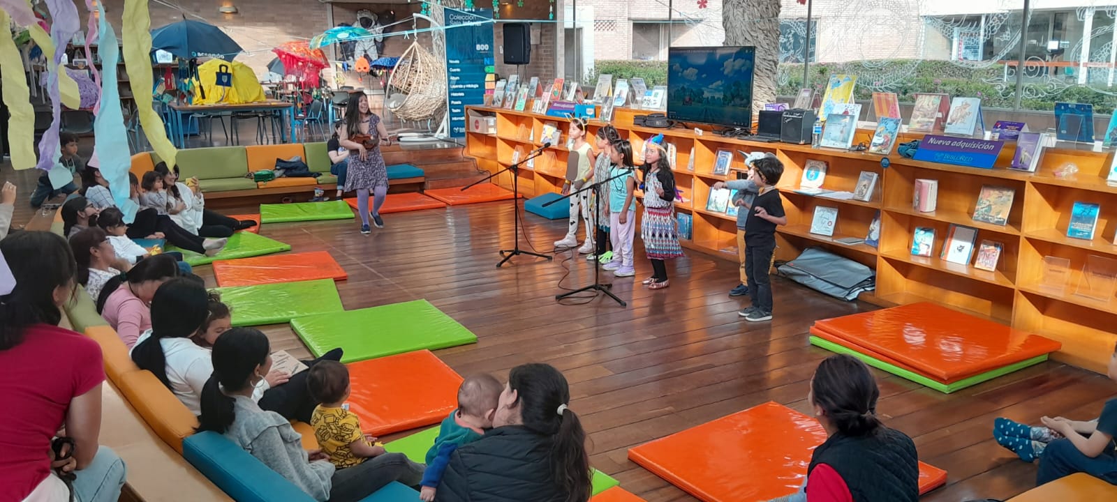 niños y niñas cantando junto a su docente frente a sus padres y cuidadores en medio del salón de una biblioteca.