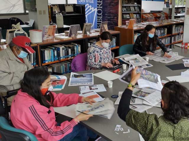 adultos y jóvenes realizando una actividad de recortar y revisar periodicos sobre un escritorio color gris.