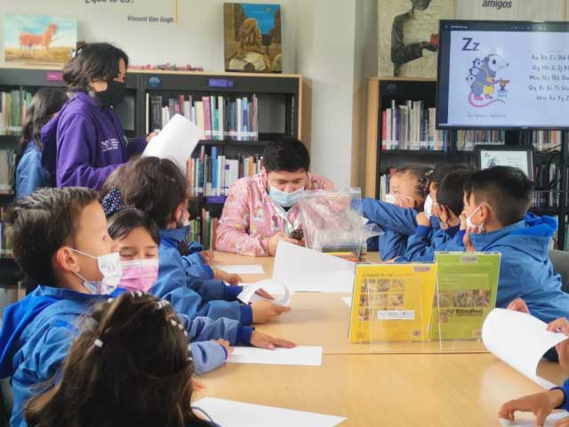 niños y niñas escribiendo sus cuentos mientras la profesora encargada del taller les lee un libro en voz alta.