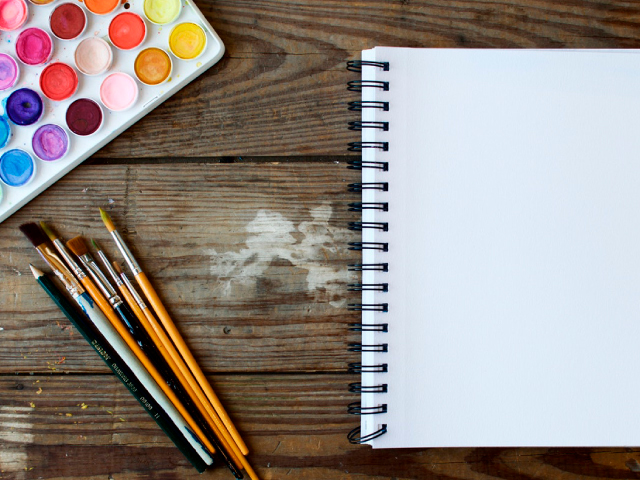 un cuaderno de hojas blancas sobre una mesa de madera junto a pinceles de varios tamaños y un set de pinturas de todos los colores.