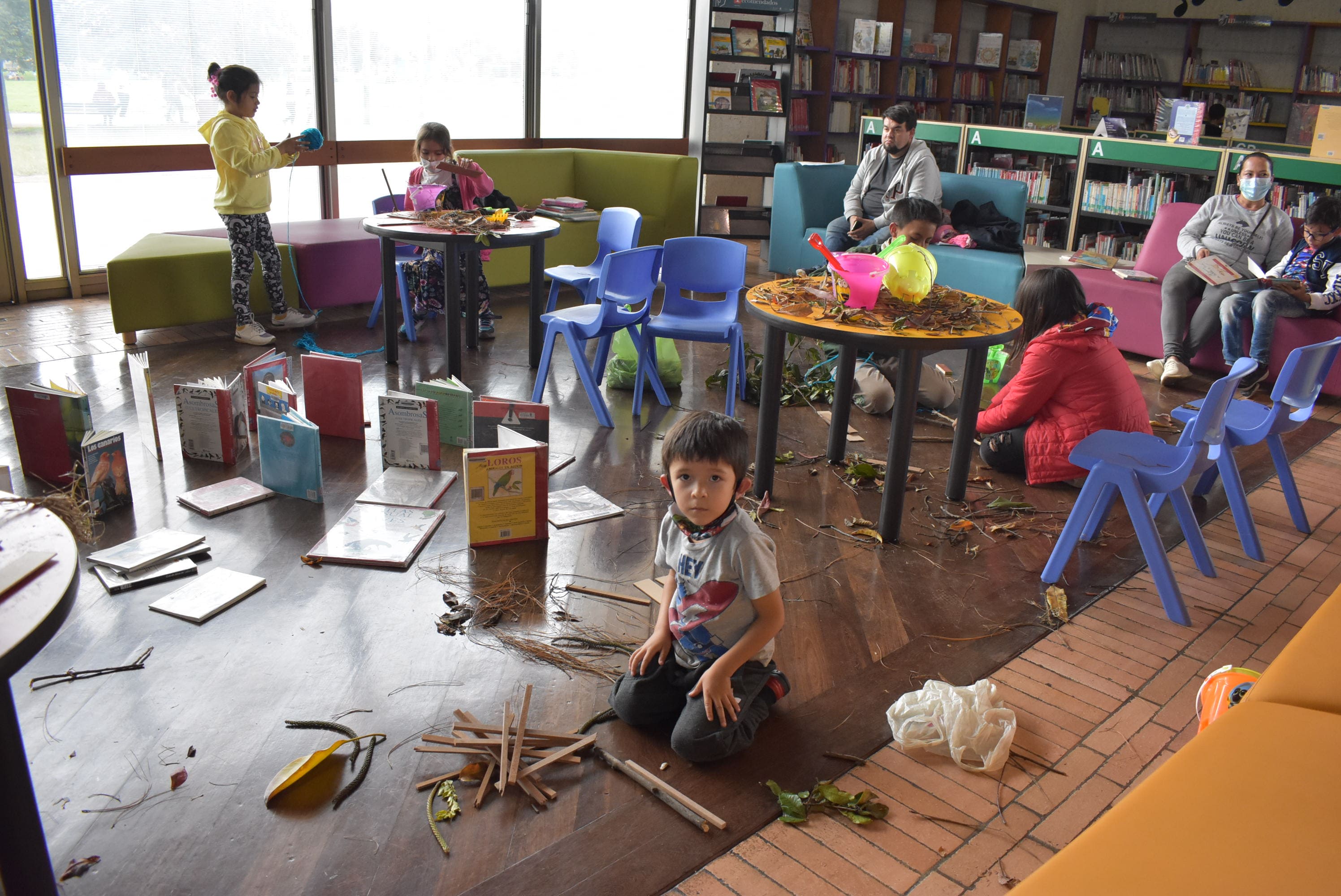 niños jugando y explorando objetos como libros, ramas, hojas, lapices, en medio del salón de una biblioteca.