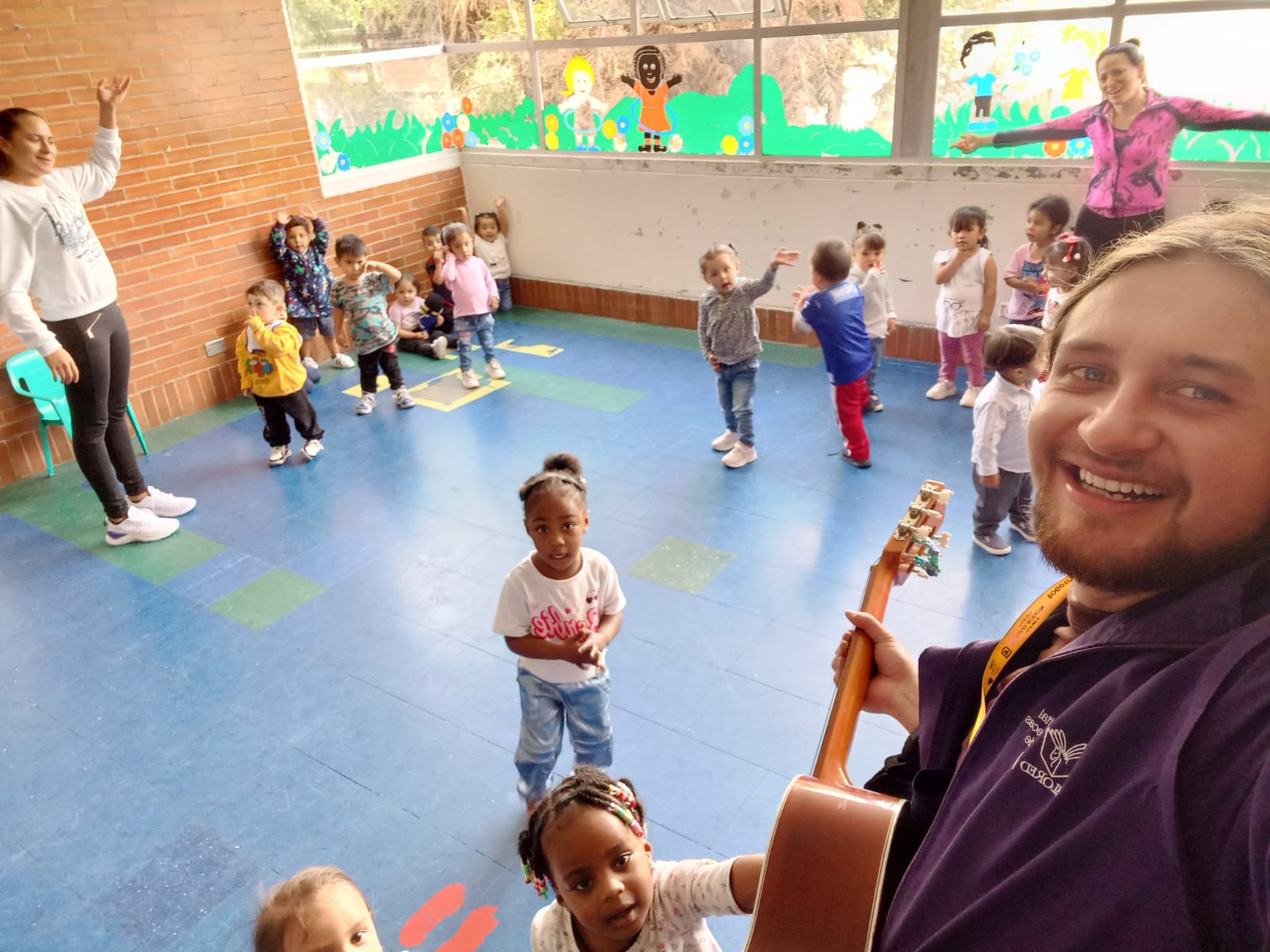 niños y niñas jugando en medio de una clase mientras su profesor toca la guitarra