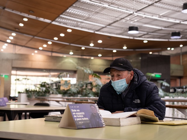 un adulto mayor leyendo un libro en medio de la sala de una biblioteca
